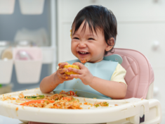 Toddler girl holding corn on a messy highchair